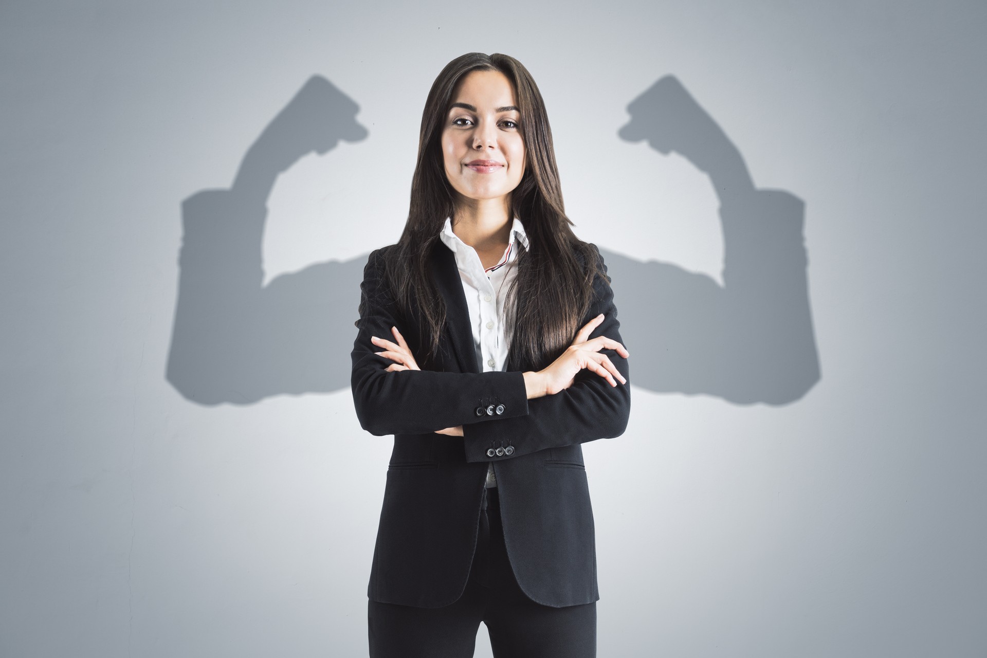 Portrait of attractive young european businesswoman with folded arms and shadow muscle arms on concrete wall background. Strenght and leadership concept.